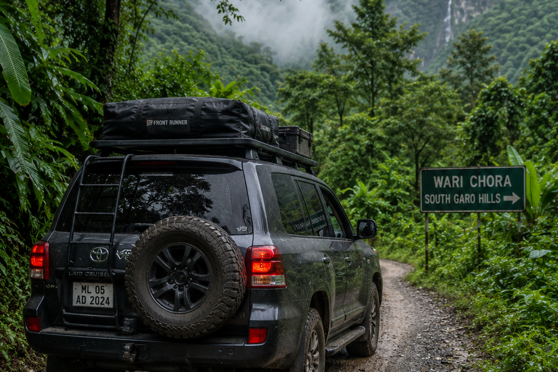 A premium 4x4 expedition vehicle driving on a remote gravel road through the lush forests of the South Garo Hills towards Wari Chora.