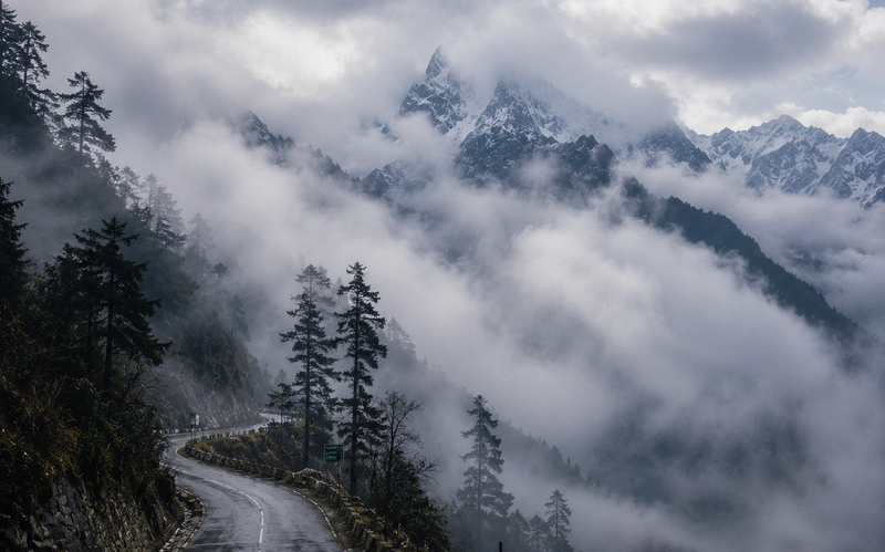 "A cinematic view of a mountain road in Sikkim being slowly enveloped by soft white clouds, with the dark silhouettes of pine trees and sharp mountain peaks peeking through the mist."