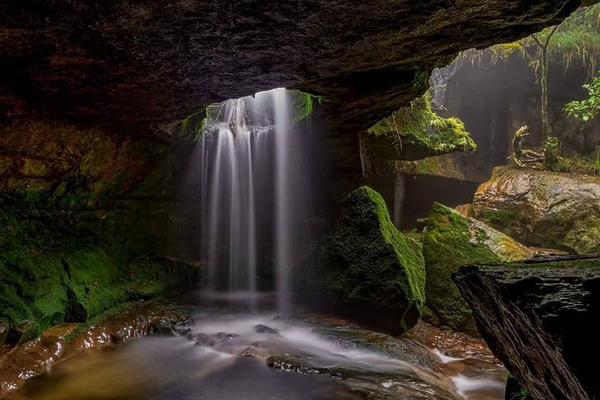 A view from inside a limestone cavern looking out at a waterfall cascading over the entrance at Garden of Caves Meghalaya.