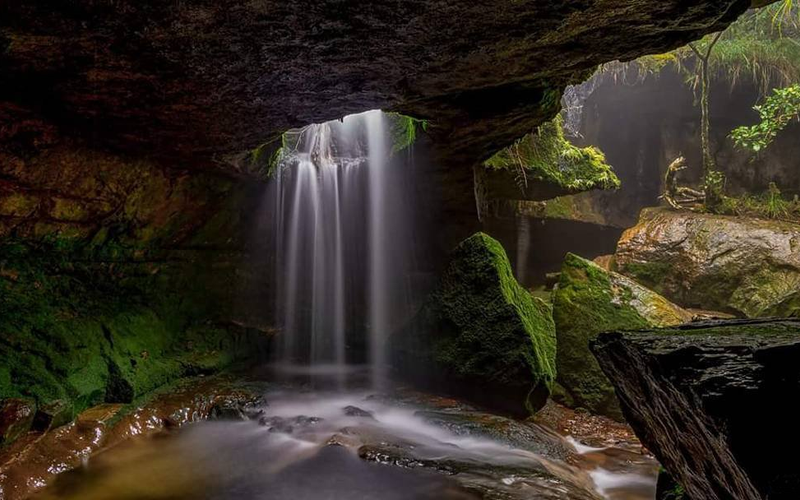 A view from inside a limestone cavern looking out at a waterfall cascading over the entrance at Garden of Caves Meghalaya.