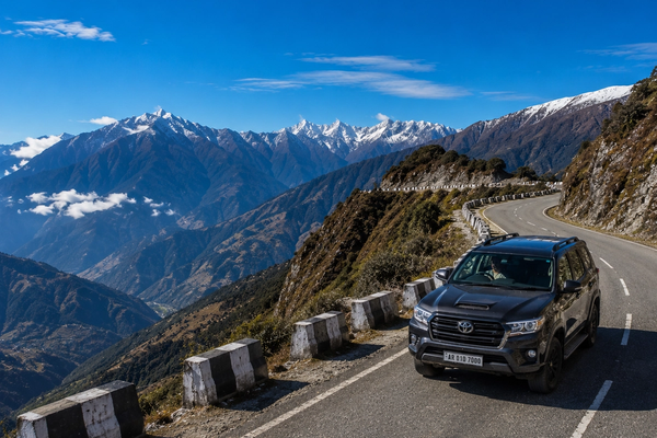 A premium 4x4 travel vehicle driving along a scenic, winding mountain highway ridge in Arunachal Pradesh under a clear blue alpine sky."
