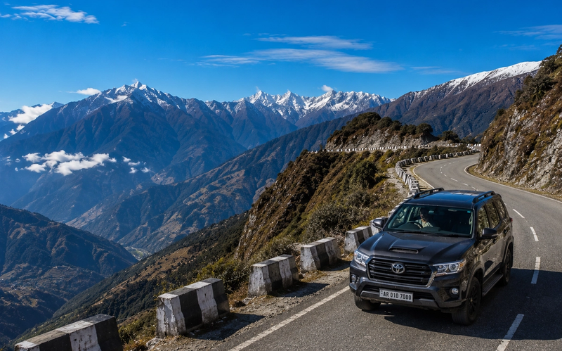 A premium 4x4 travel vehicle driving along a scenic, winding mountain highway ridge in Arunachal Pradesh under a clear blue alpine sky."