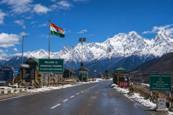 A cinematic, panoramic shot of a scenic mountain highway checkpost in Arunachal Pradesh under a clear alpine sky with snow-capped peaks in the background.
