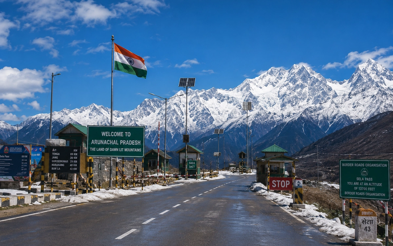 A cinematic, panoramic shot of a scenic mountain highway checkpost in Arunachal Pradesh under a clear alpine sky with snow-capped peaks in the background.