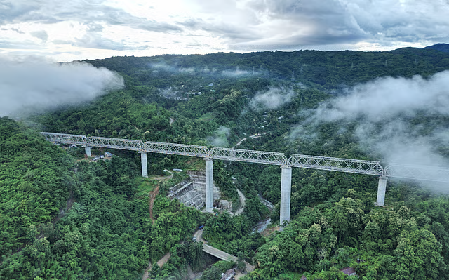 Passenger train crossing a tall concrete bridge over forested hills on the Bairabi Sairang railway line in Mizoram