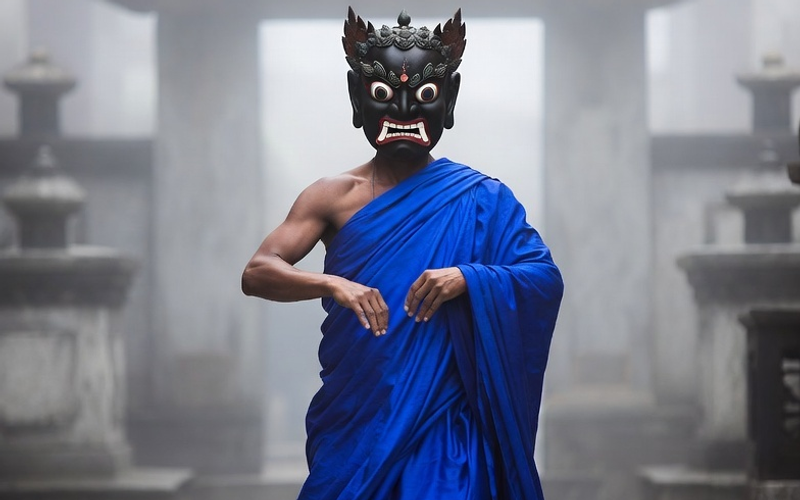"A monk wearing a vibrant blue silk robe and a traditional carved wrathful deity mask performing the sacred Cham dance in a misty monastery courtyard in Sikkim."