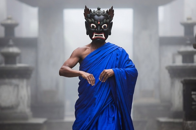 "A monk wearing a vibrant blue silk robe and a traditional carved wrathful deity mask performing the sacred Cham dance in a misty monastery courtyard in Sikkim."
