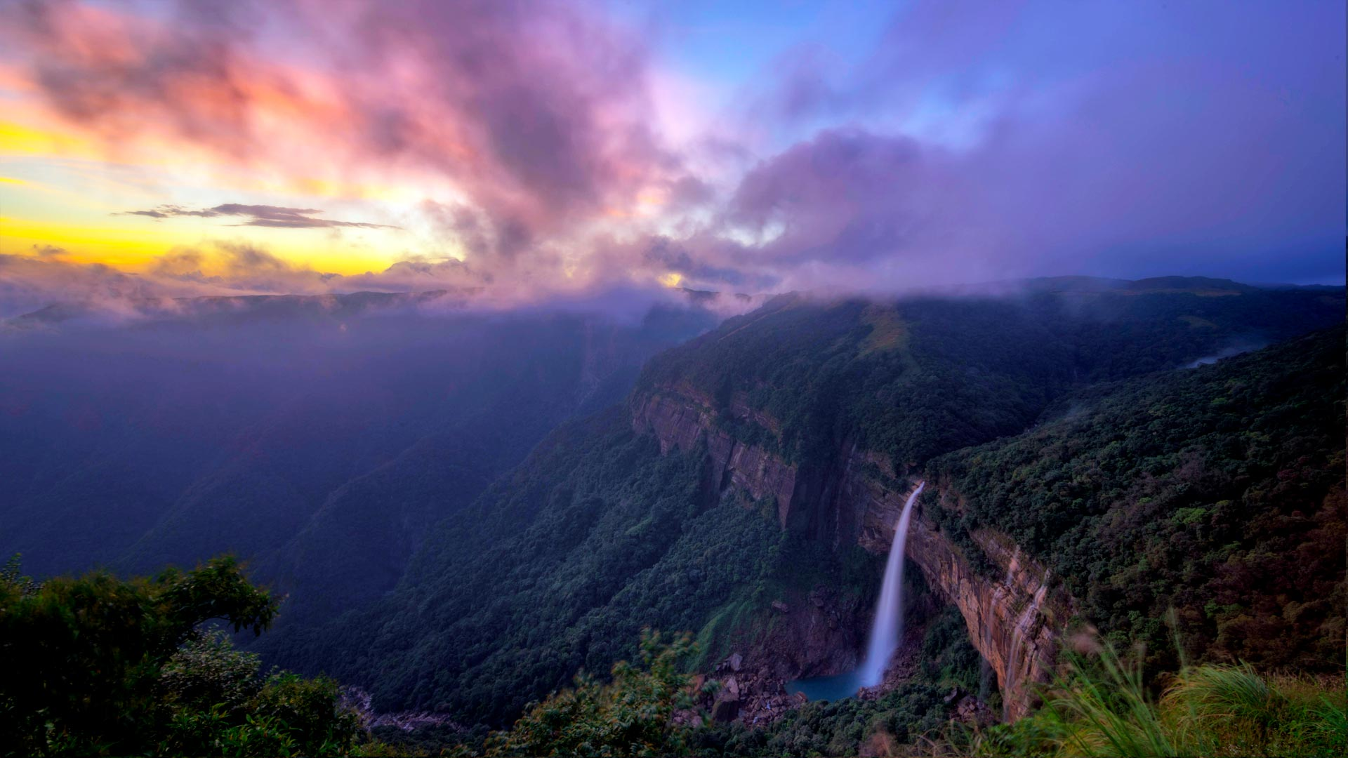 A wide vertical shot of Nohkalikai Falls in Meghalaya, showing the tall white waterfall plunging from a lush green plateau into a turquoise blue pool below, surrounded by dense tropical forest and mist.