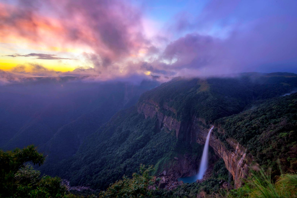 A wide vertical shot of Nohkalikai Falls in Meghalaya, showing the tall white waterfall plunging from a lush green plateau into a turquoise blue pool below, surrounded by dense tropical forest and mist.