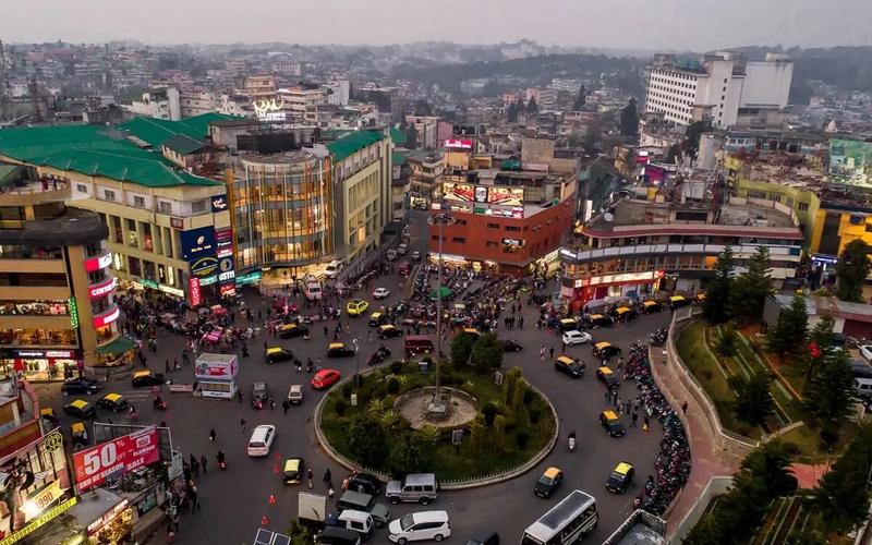 Aerial view of the Police Bazar roundabout in Shillong, Meghalaya, showing the iconic central circle and commercial buildings like the M-Crown Hotel at dusk.