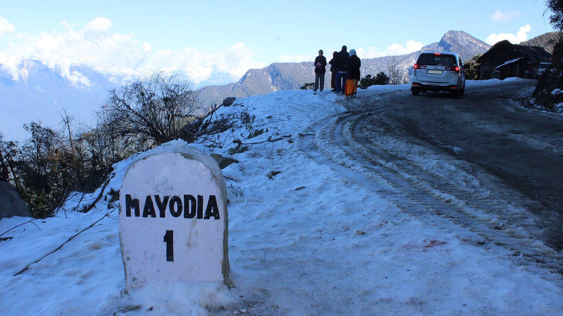 A Mayodia milestone half buried in snow on the road near Mayudia Pass, with a car and snowy hills behind