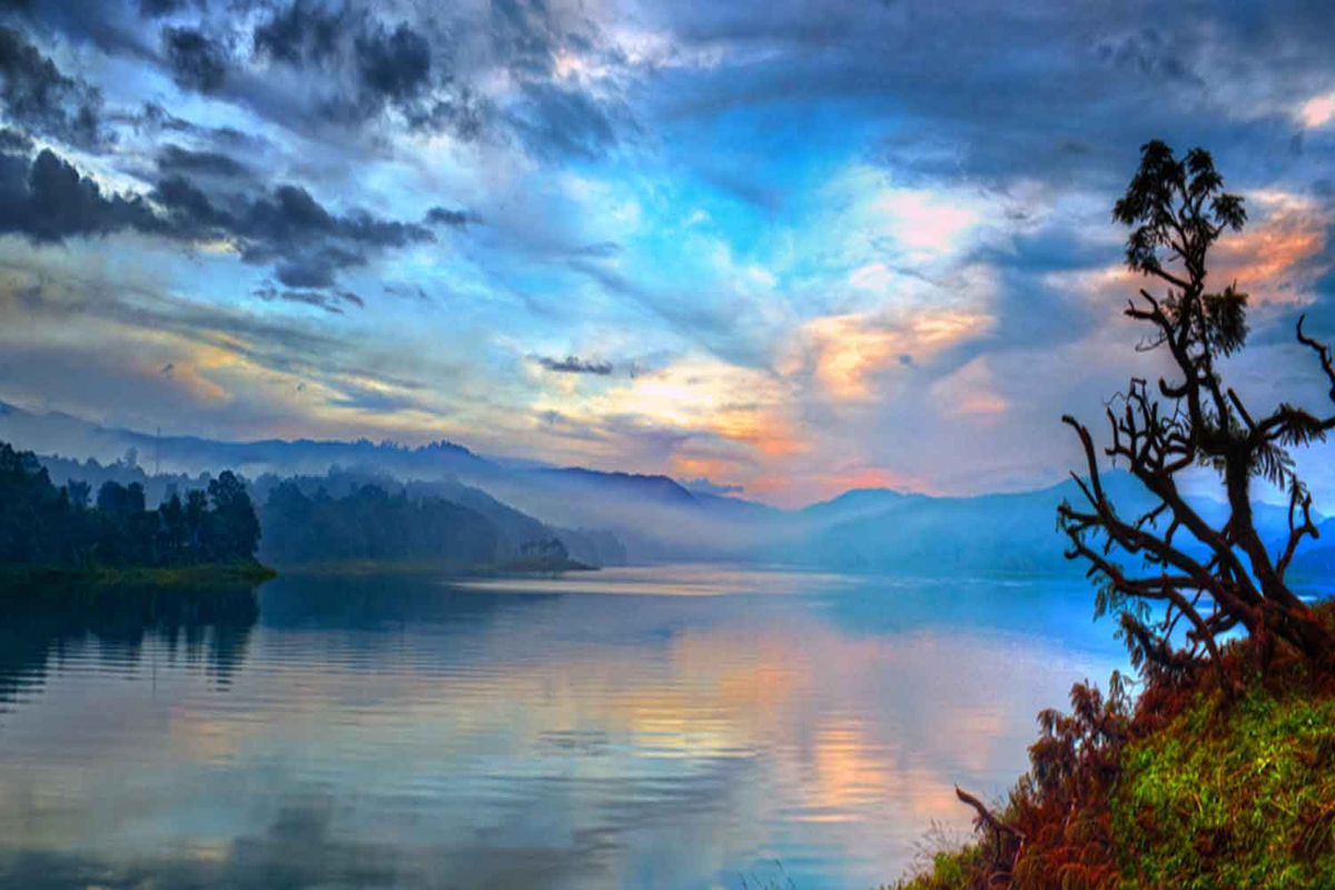 Umiam Lake Shillong at sunrise with calm emerald waters reflecting a dramatic blue and orange sky, misty hills and silhouetted trees in Meghalaya