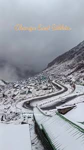 "A wide-angle panoramic view of the winding mountain roads leading to Nathu La Pass in East Sikkim, featuring sharp hairpin turns and snow-dusted peaks under a clear blue sky."