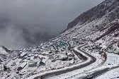 "A wide-angle panoramic view of the winding mountain roads leading to Nathu La Pass in East Sikkim, featuring sharp hairpin turns and snow-dusted peaks under a clear blue sky."