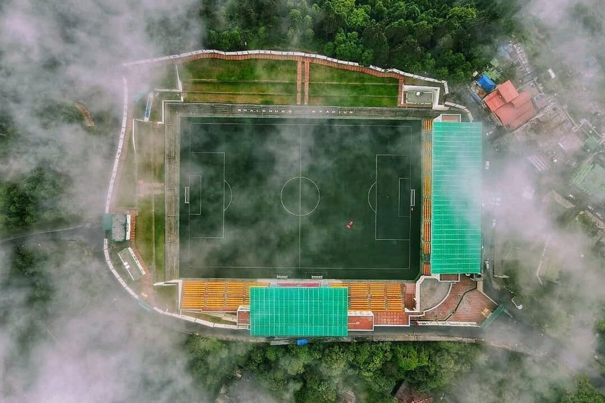 A high-altitude green football turf at Baichung Stadium in Namchi with hills and residential houses in the background.