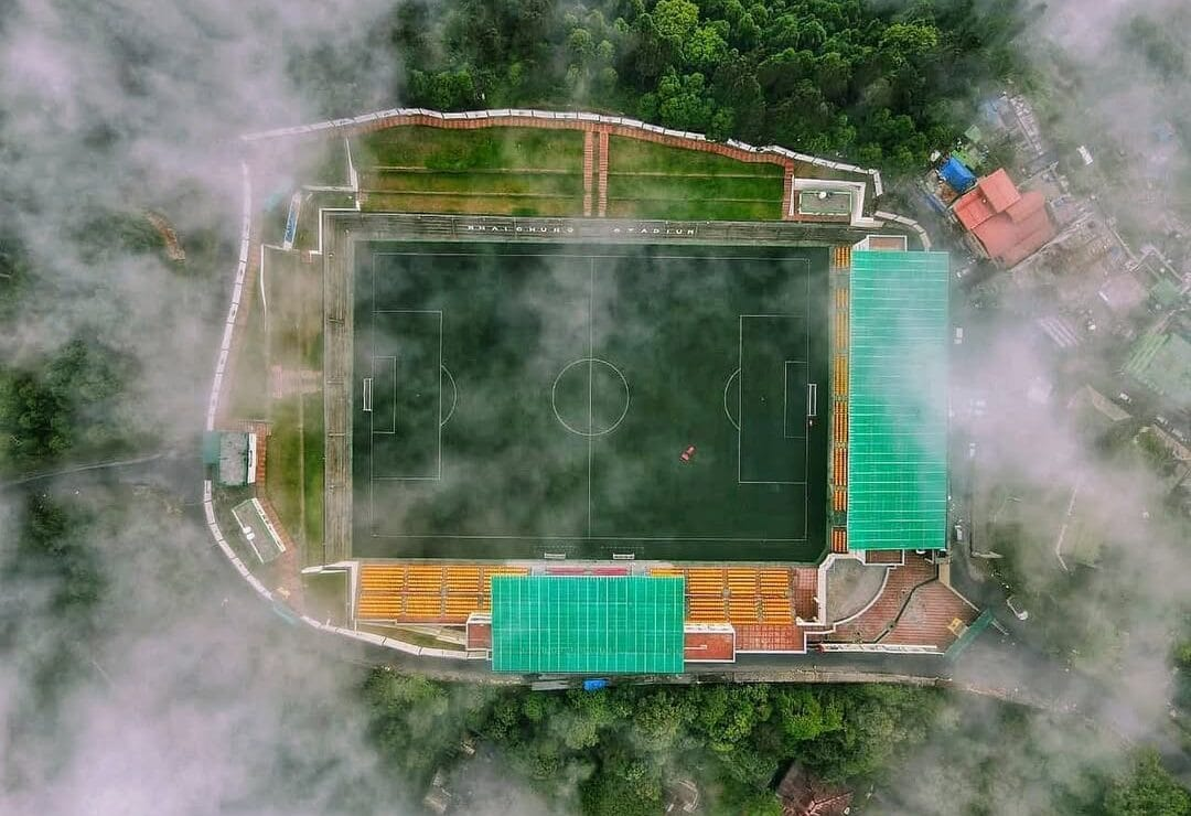 A high-altitude green football turf at Baichung Stadium in Namchi with hills and residential houses in the background.