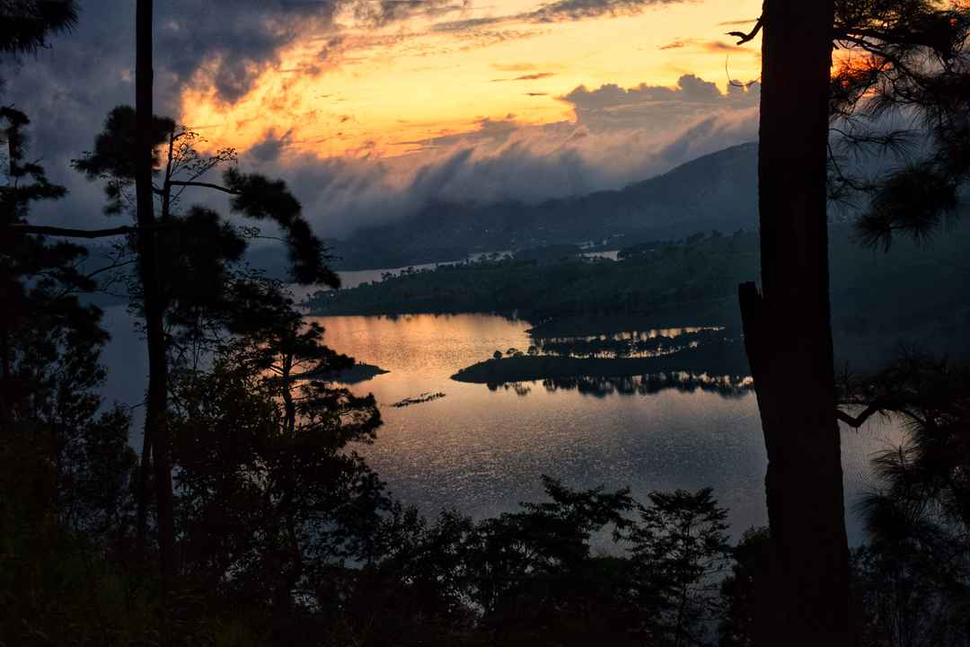 A wide view of Umiam Lake in Meghalaya during sunset, showing serene blue waters and surrounding pine forests.