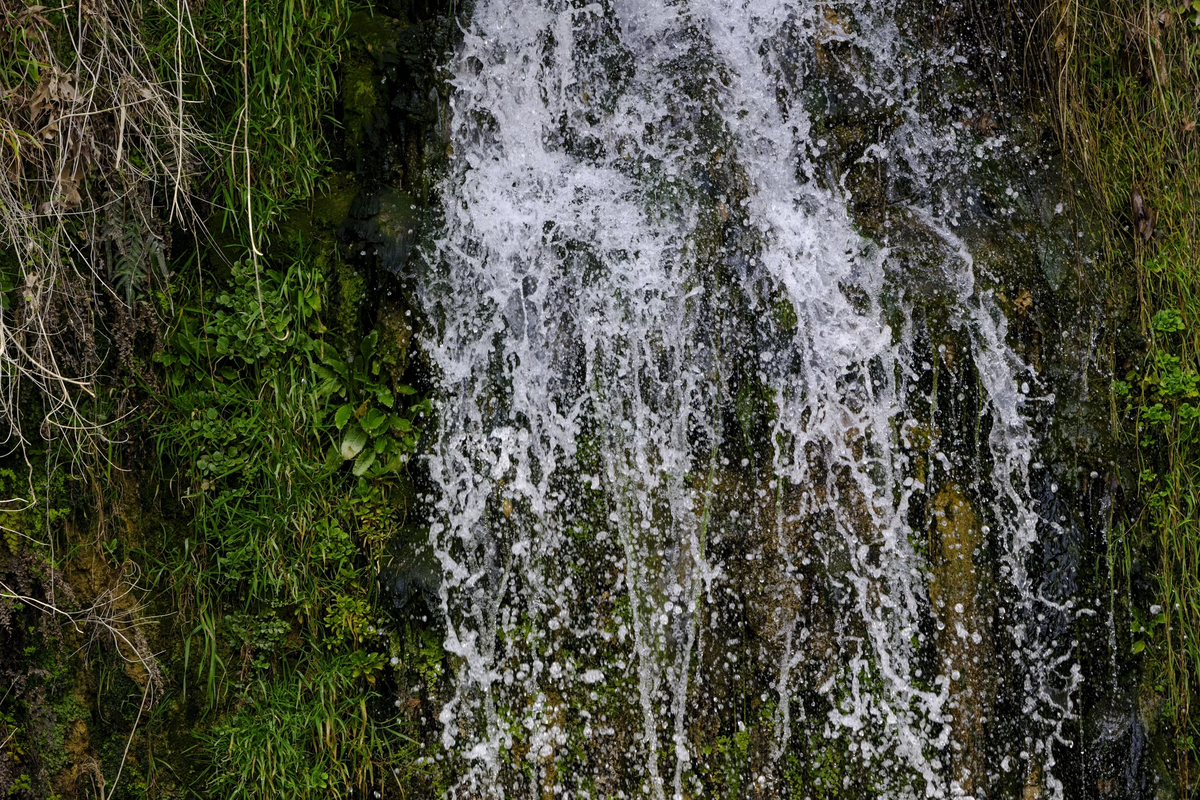 A small waterfall cascading down a tall green mossy cliff into a narrow river channel.