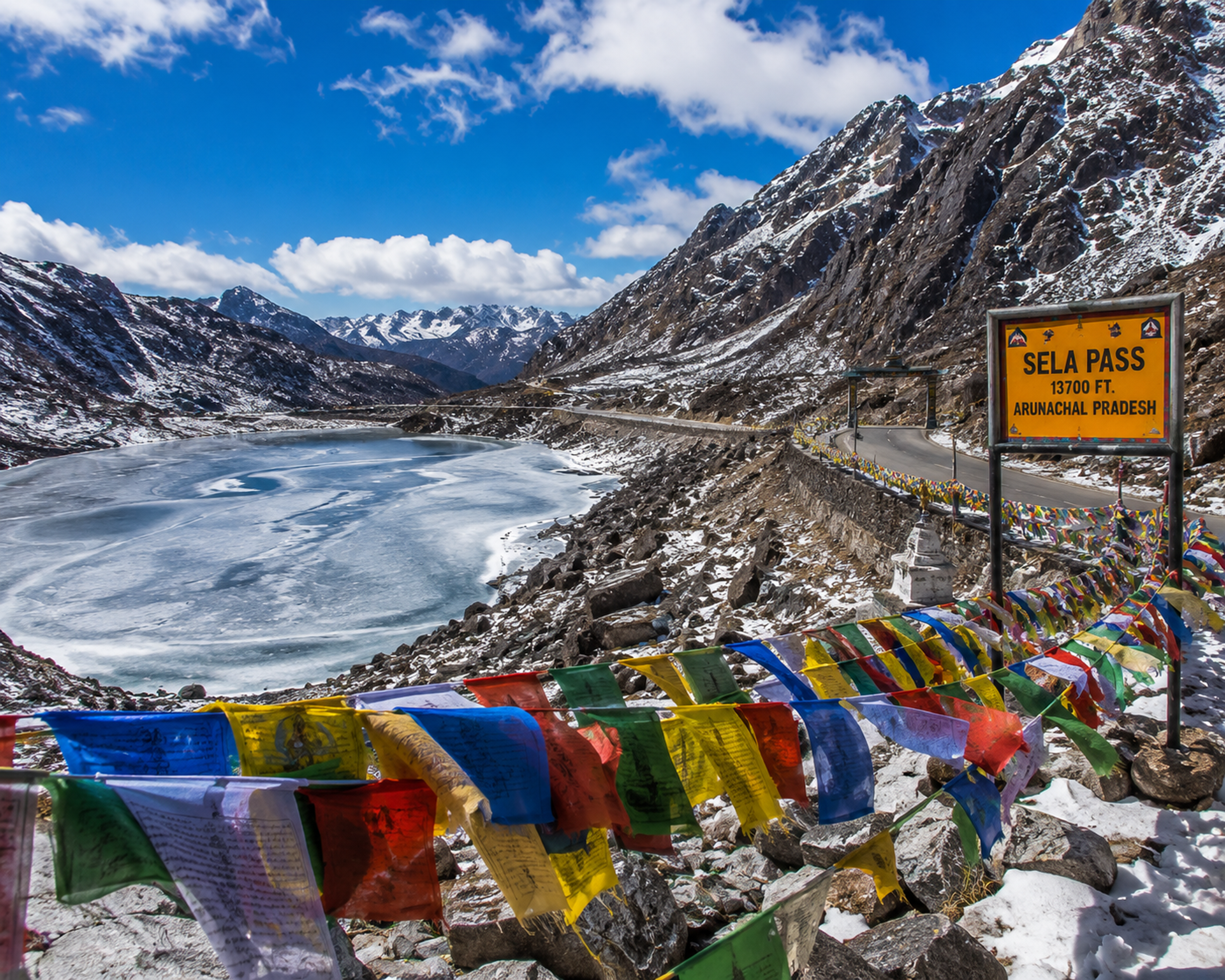 A high alpine road looping past a frozen mountain lake with colorful prayer flags at Sela Pass in Arunachal Pradesh.