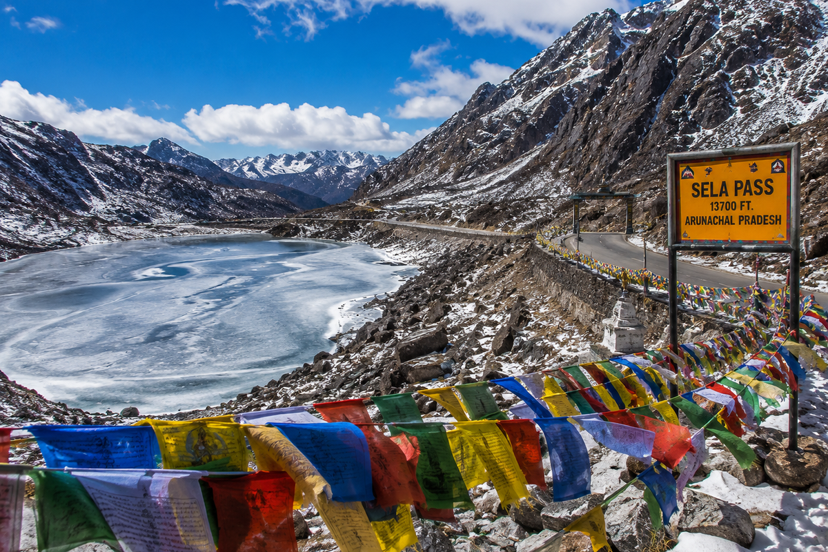 A high alpine road looping past a frozen mountain lake with colorful prayer flags at Sela Pass in Arunachal Pradesh.