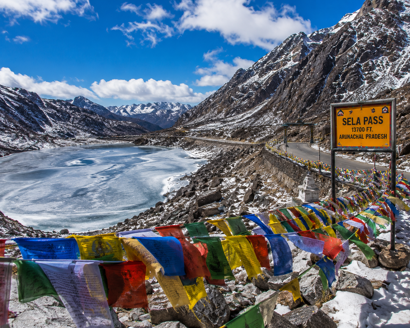 A high alpine road looping past a frozen mountain lake with colorful prayer flags at Sela Pass in Arunachal Pradesh.
