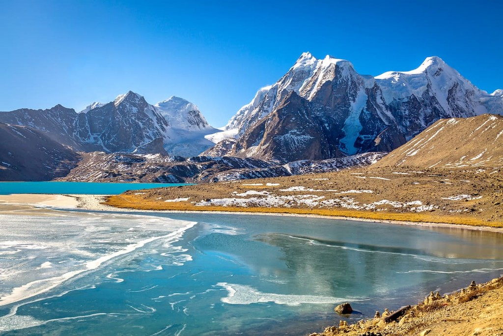 The crystal-clear turquoise waters of Gurudongmar Lake, with prayer flags in the foreground and the snow-capped Himalayan peaks of the Tibetan Plateau in the background