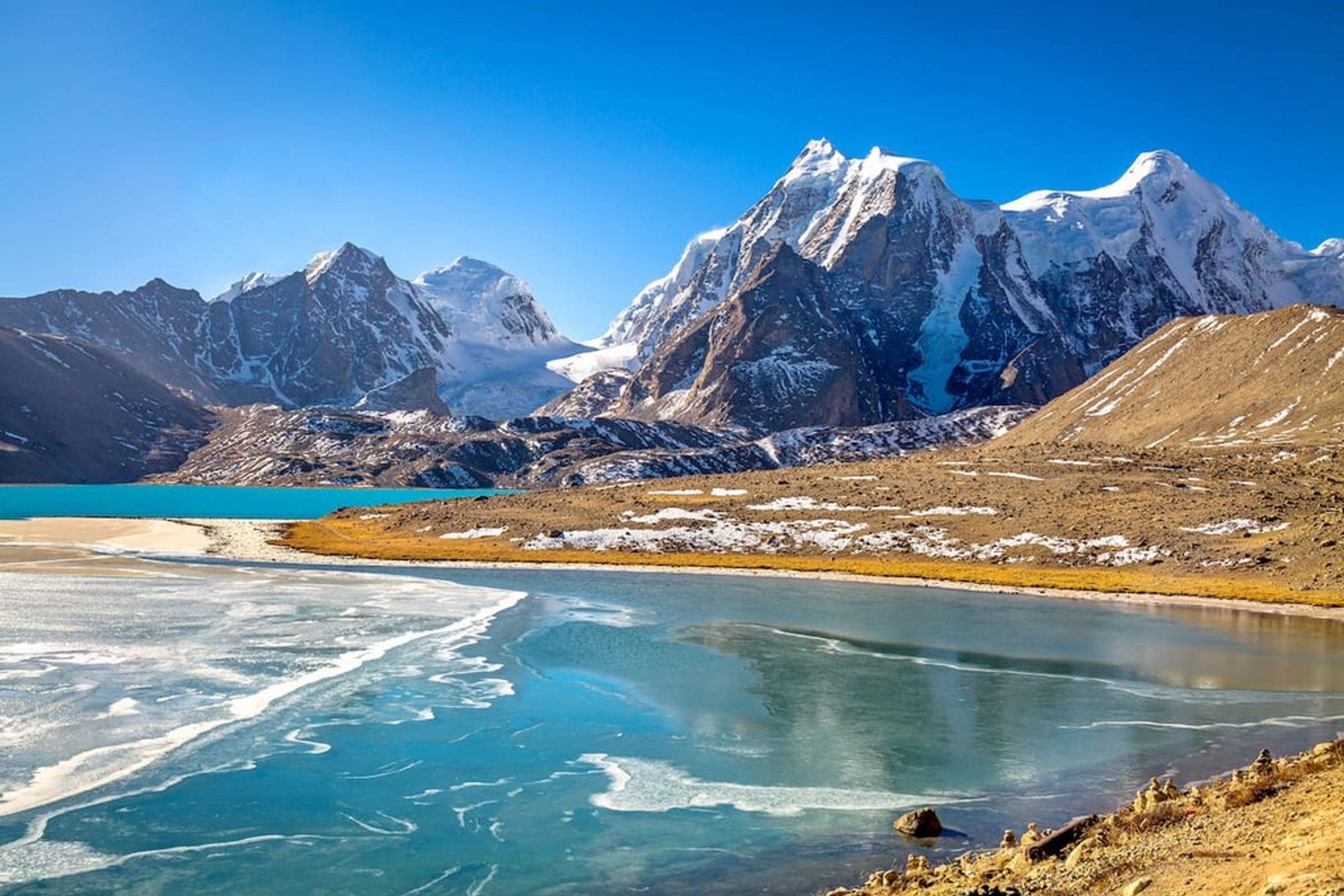The crystal-clear turquoise waters of Gurudongmar Lake, with prayer flags in the foreground and the snow-capped Himalayan peaks of the Tibetan Plateau in the background