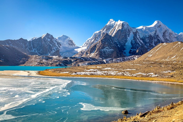 The crystal-clear turquoise waters of Gurudongmar Lake, with prayer flags in the foreground and the snow-capped Himalayan peaks of the Tibetan Plateau in the background