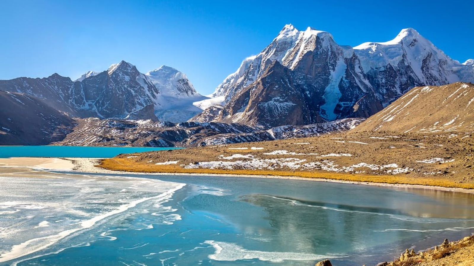 The crystal-clear turquoise waters of Gurudongmar Lake, with prayer flags in the foreground and the snow-capped Himalayan peaks of the Tibetan Plateau in the background