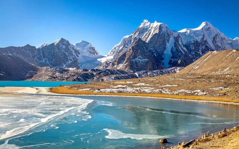 The crystal-clear turquoise waters of Gurudongmar Lake, with prayer flags in the foreground and the snow-capped Himalayan peaks of the Tibetan Plateau in the background