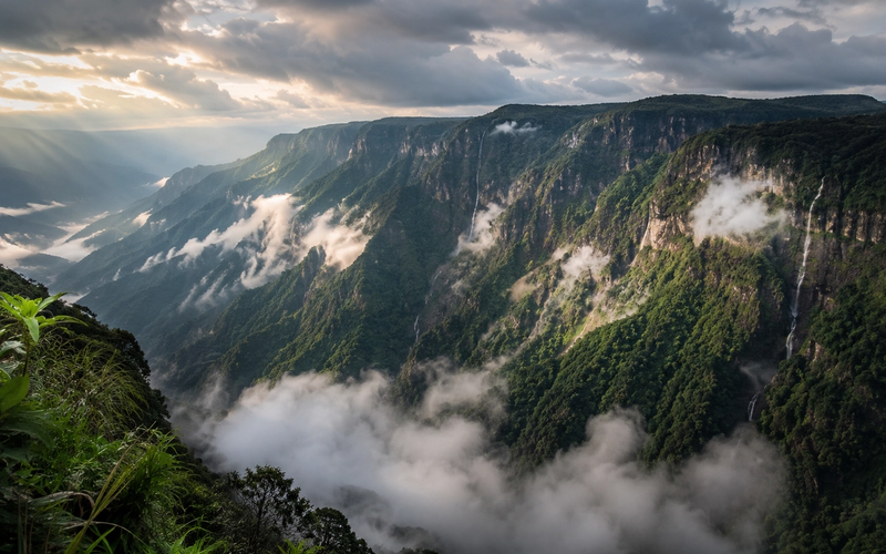 A cinematic panoramic view of a deep mountain gorge in Cherrapunji, Meghalaya, with dramatic mists and clouds parting over emerald-green cliffs."
