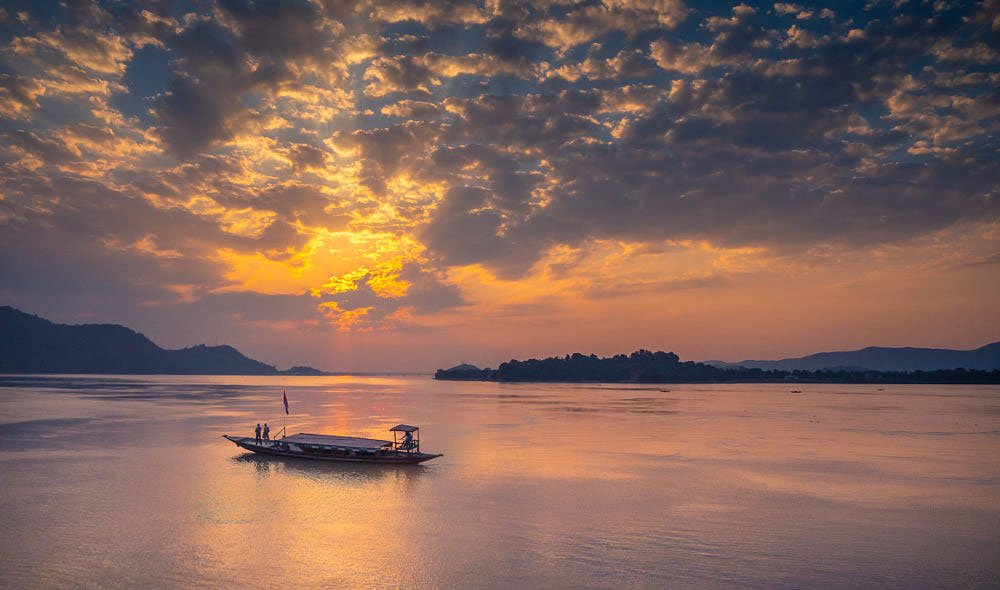 A traditional wooden ferry boat carrying passengers across the Brahmaputra River in Guwahati at sunset, with the silhouette of Umananda Island and its temple in the distance under a golden orange sky.