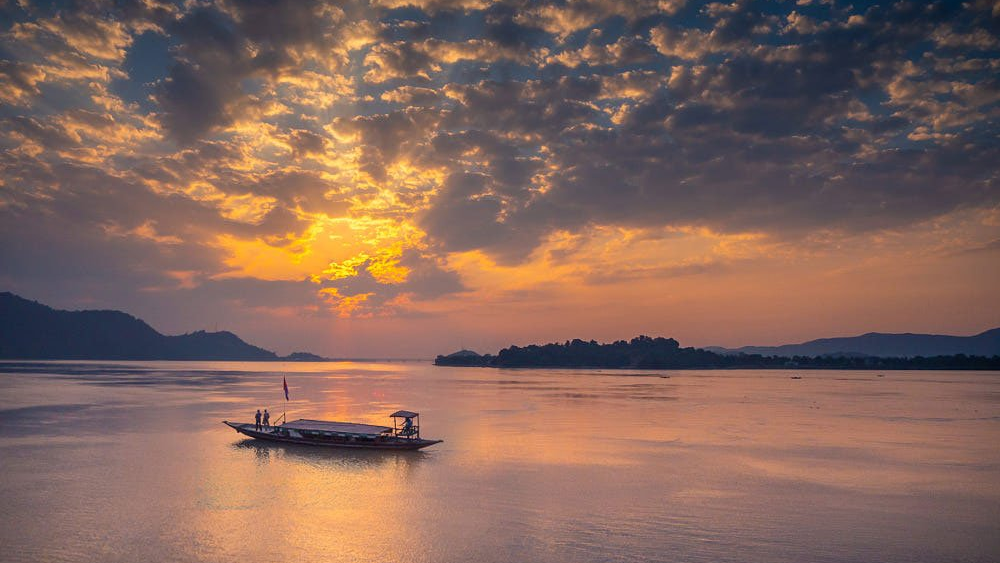A traditional wooden ferry boat carrying passengers across the Brahmaputra River in Guwahati at sunset, with the silhouette of Umananda Island and its temple in the distance under a golden orange sky.