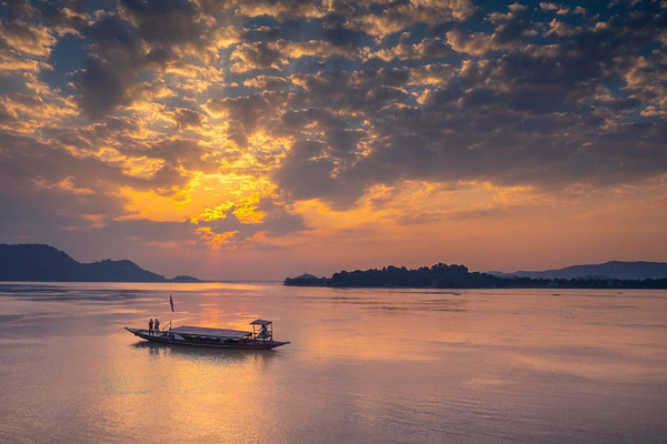 A traditional wooden ferry boat carrying passengers across the Brahmaputra River in Guwahati at sunset, with the silhouette of Umananda Island and its temple in the distance under a golden orange sky.