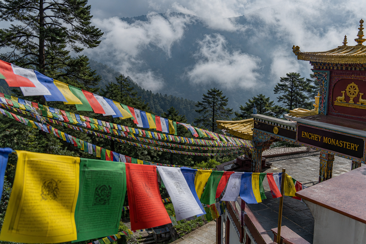 Colorful Buddhist prayer flags outside Enchey Monastery in Gangtok, with the misty Himalayan landscape and pine forests in the background.