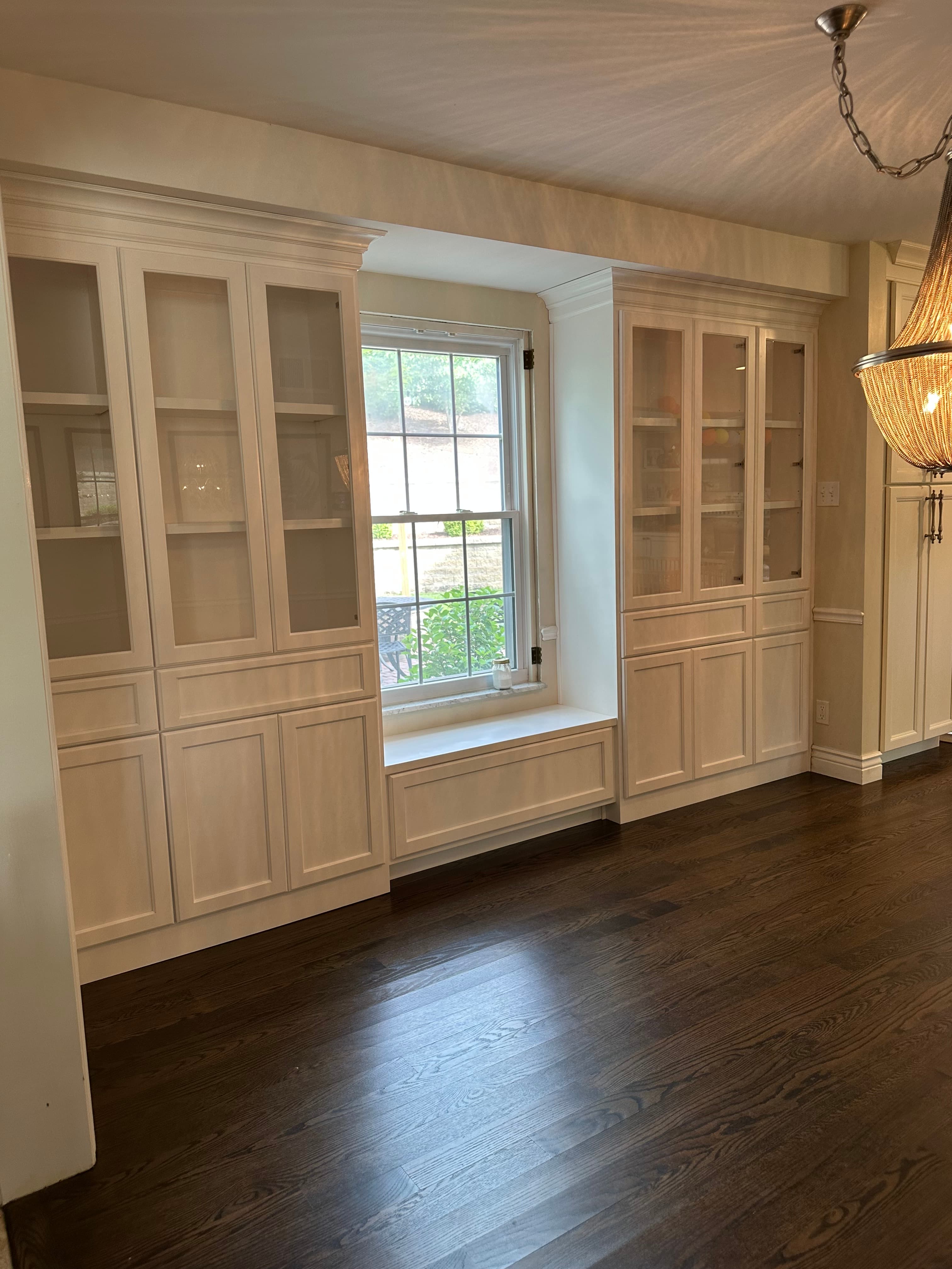 White floor-to-ceiling built-in cabinets with glass doors surrounding a window in a finished room