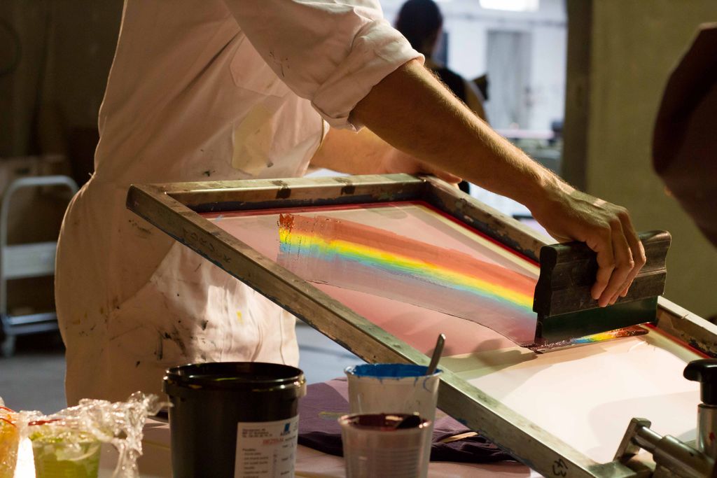 Barrierefreiheit im Kopf 2015: Siebdruck-Workshop: Eine Hand zieht Regenbogenfarbe mit dem Rakel über das Sieb