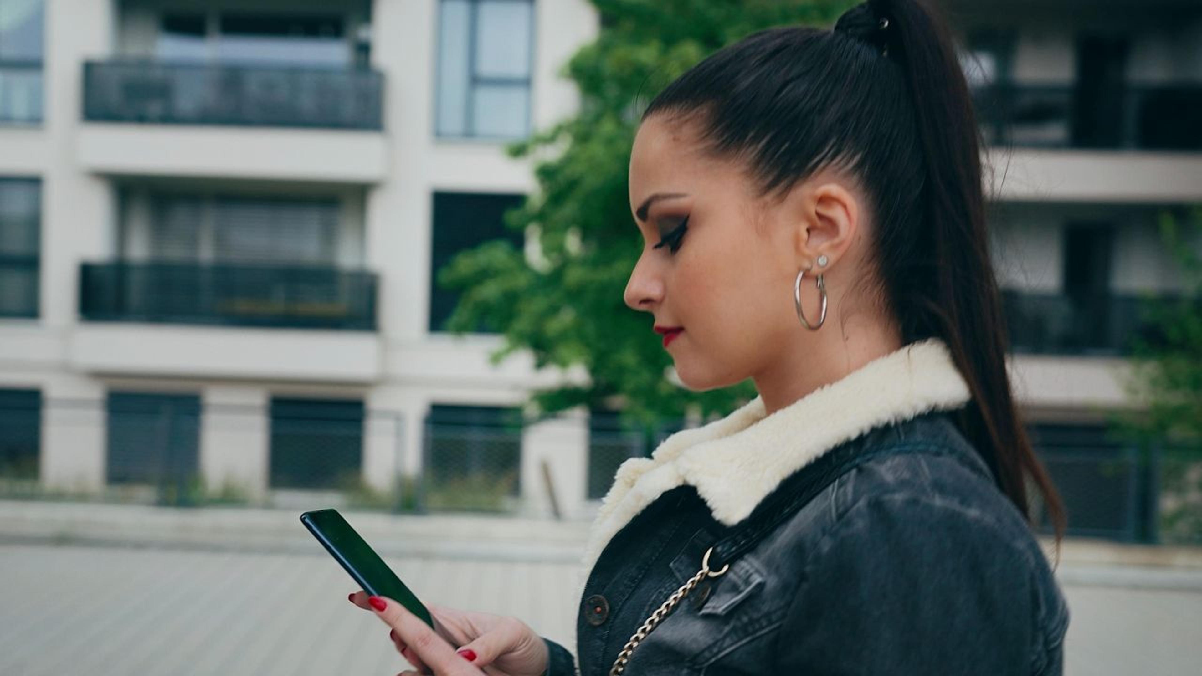 Frau mit schwarzen Haaren, geht draußen rum und guckt auf ihr Handy