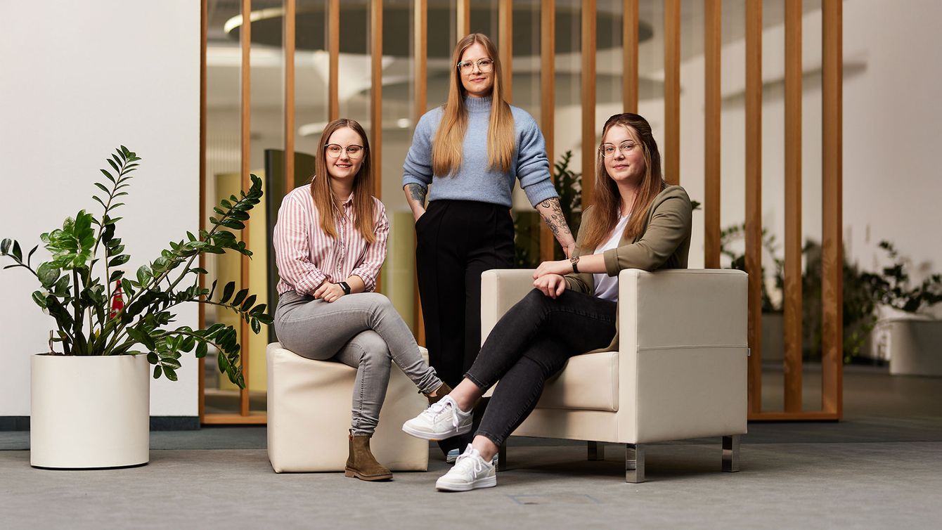 Drei junge Frauen, zwei sitzen, eine steht zwischen ihnen. 