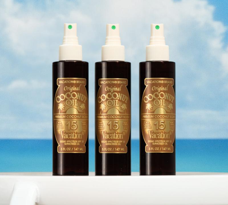 Three Vacation Brand Original Coconut Oil SPF 15 sunscreen spray bottles against a blue sky and ocean.