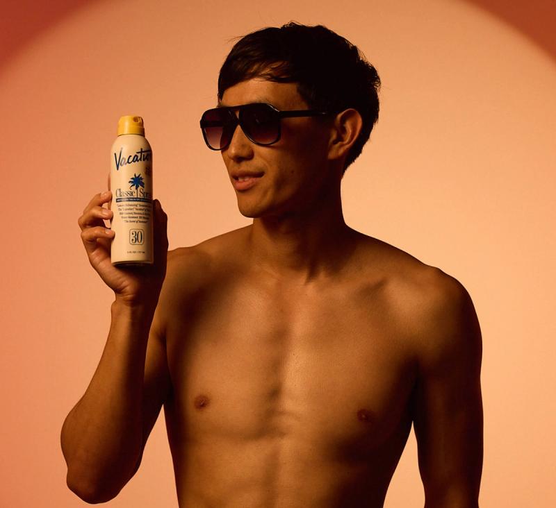 A shirtless man in sunglasses holds up a bottle of Vacation Classic Spray sunscreen.