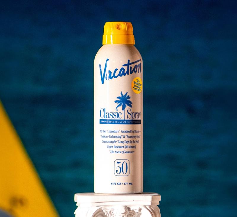 Vacation Classic Spray SPF 50 sunscreen can with a yellow cap and palm tree logo, on a pedestal against a blue background.