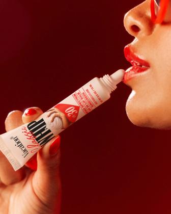A hand with red nails applies Vacation Classic WHIP SPF 30 Lip Balm to red lips.