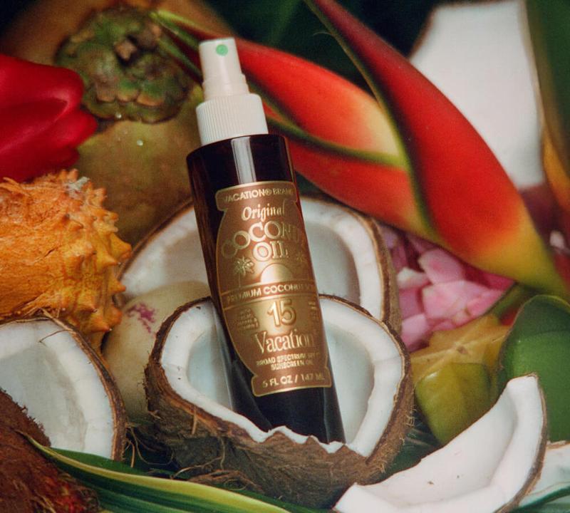 A bottle of Vacation Brand Original Coconut Oil with SPF 15, surrounded by coconut halves, tropical fruits, and red flowers.