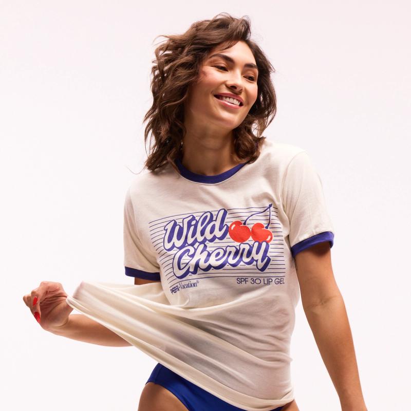 A smiling woman wearing a white ringer tee with a "Wild Cherry" graphic and blue bottoms.