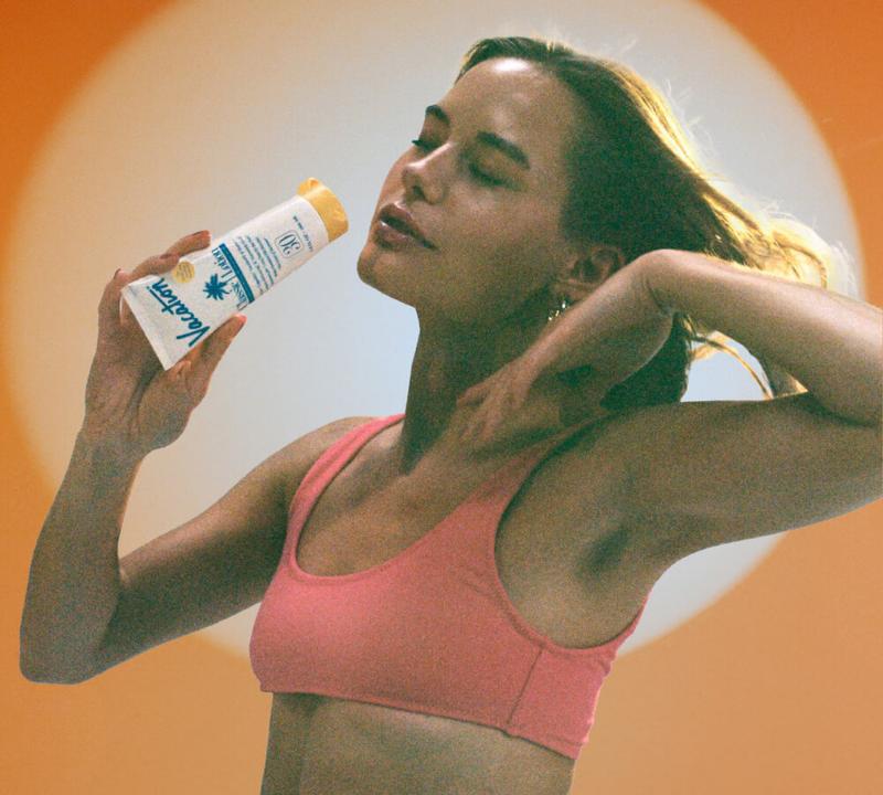A woman in a pink top smells a "Vacation" sunscreen tube with her eyes closed, against an orange and light background.