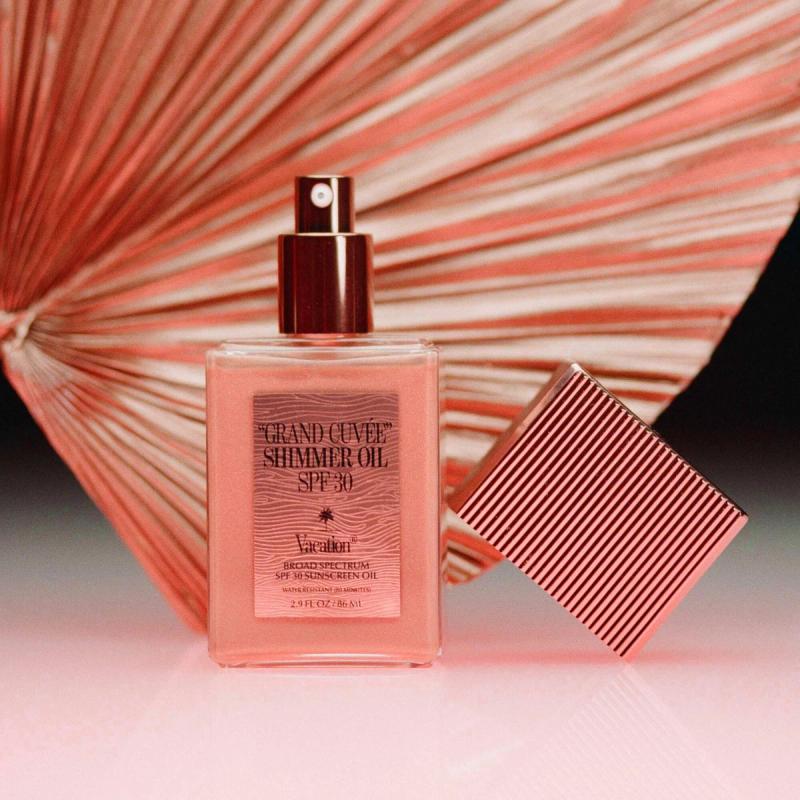 A clear bottle of pink "Vacation Grand Cuvée Shimmer Oil SPF 30" with its ribbed cap beside it, set against a coral fan.