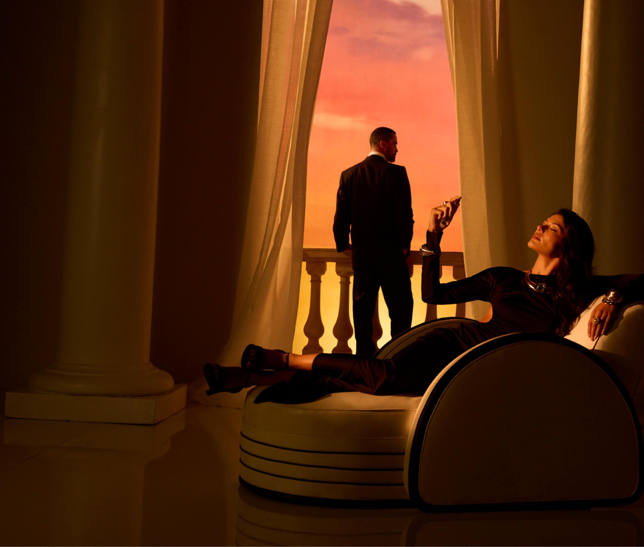 A man in a suit stands on a balcony facing a vibrant sunset, while a woman reclines on an elegant indoor chair, holding an object.
