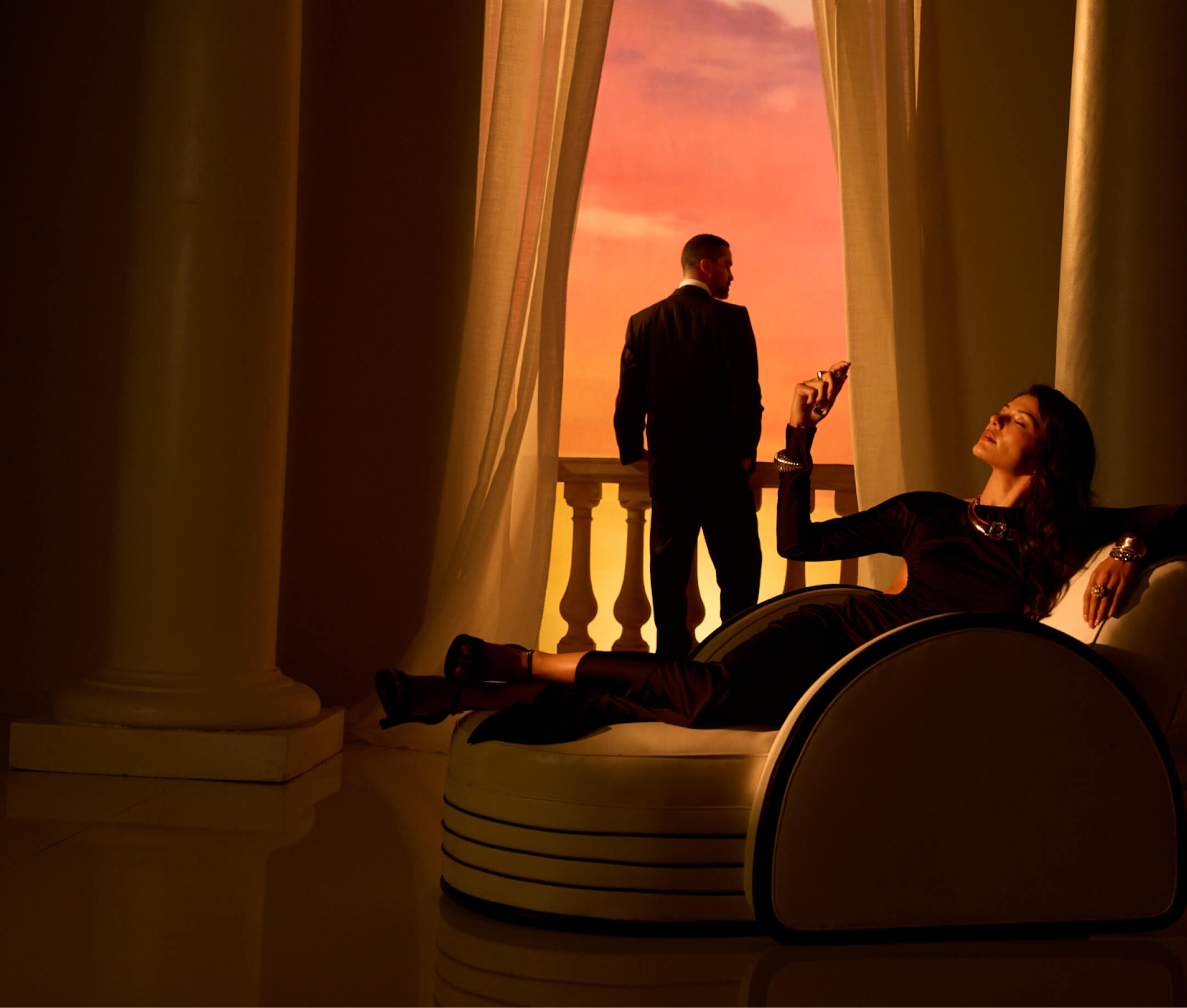 A man in a suit stands on a balcony facing a vibrant sunset, while a woman reclines on an elegant indoor chair, holding an object.