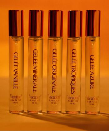 Five clear glass Vacation rollerball fragrance bottles with gold caps stand in a row against an orange background.